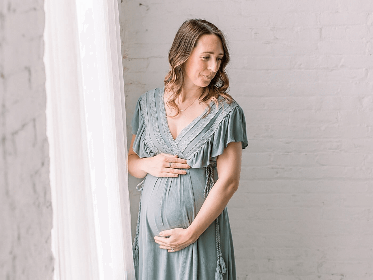 Pregnant woman wearing a sage green dress for maternity photos in a natural light studio. She is standing in front of a window.