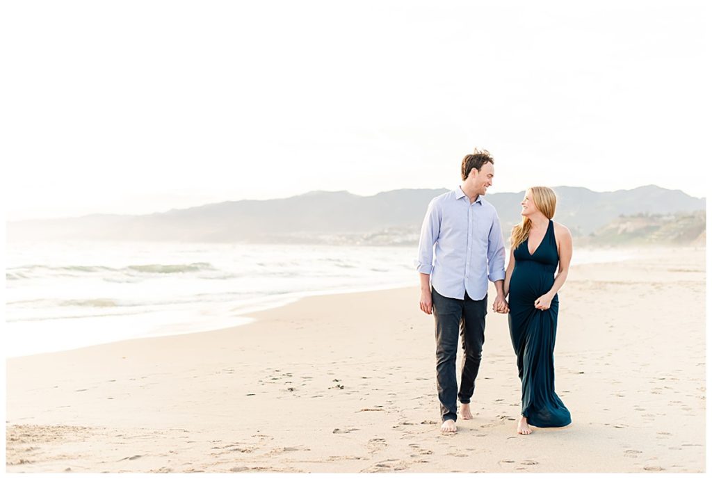 Image shows a couple at their maternity session on the beach walking together while holding hands and smiling at eachother. 