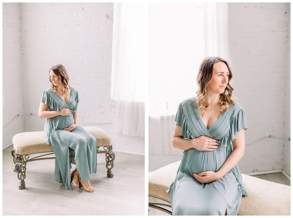 Image shows a pregnant woman sitting on a decorative bench wearing a sage green dress for her maternity photos in a natural light studio in New Jersey