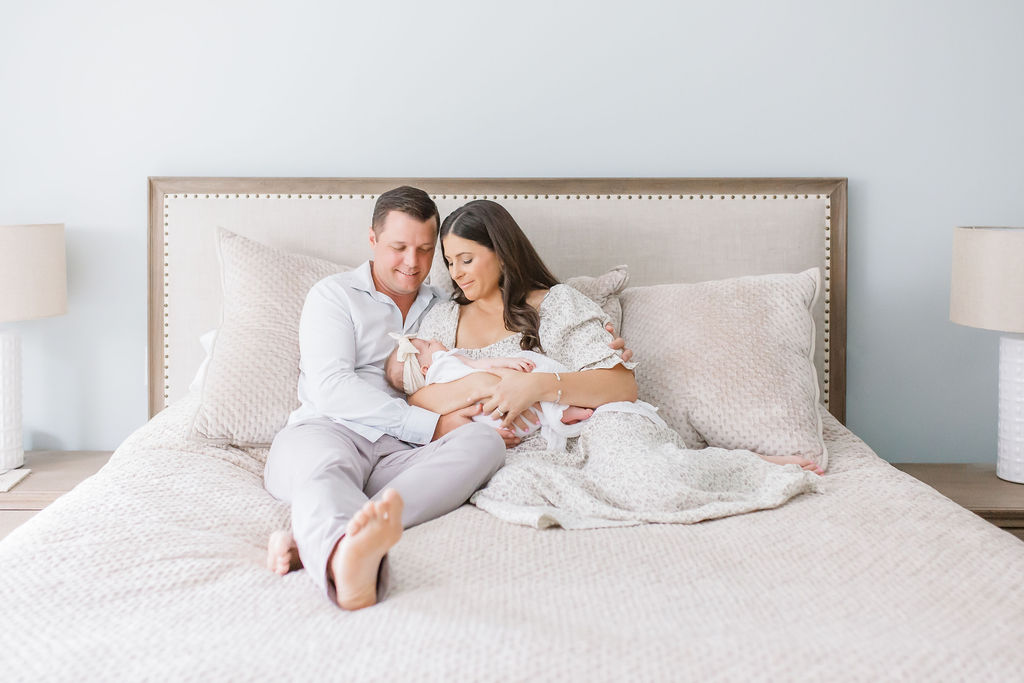 A mother and father sit together on their bed while cradling their newborn baby