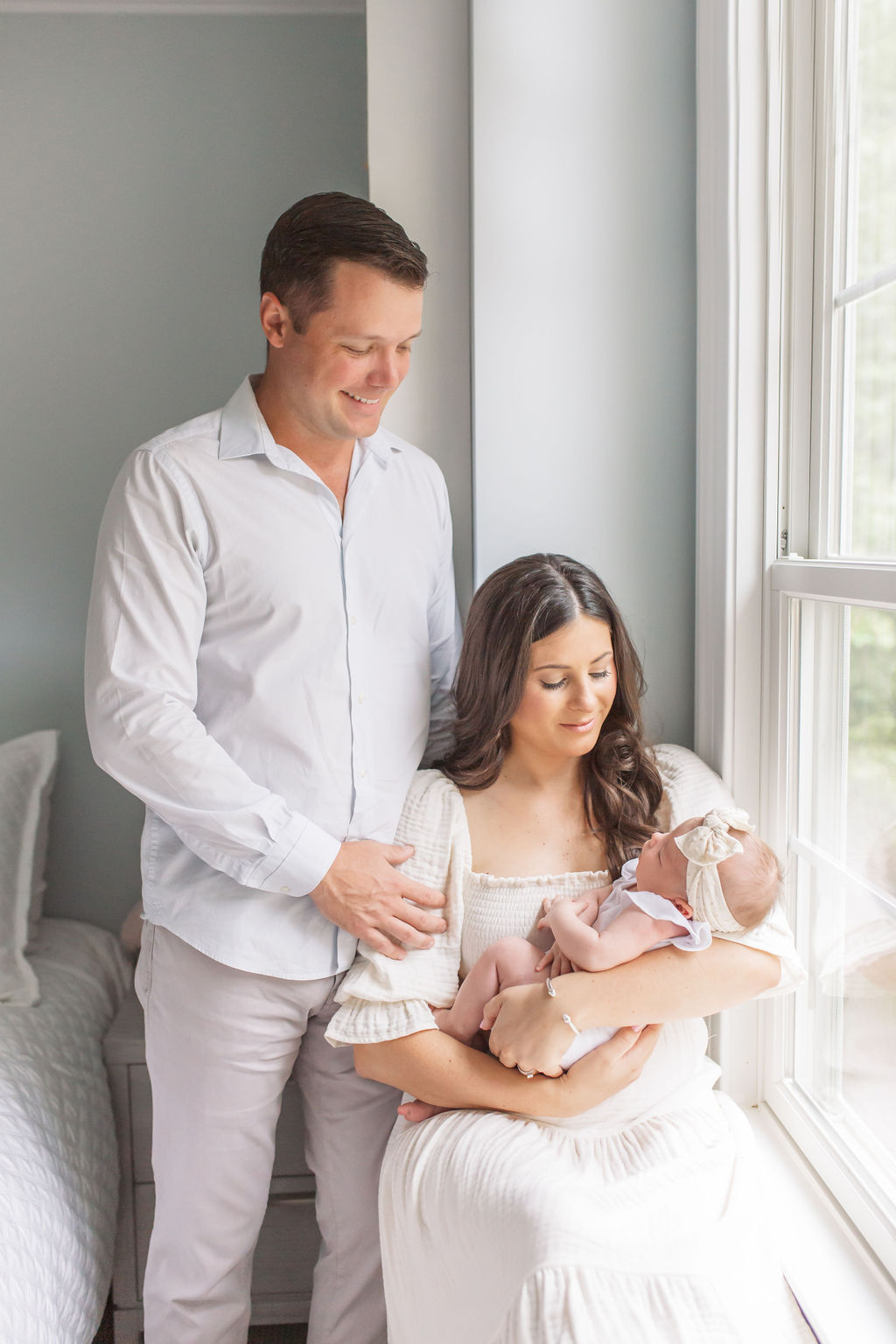 A husband and wife hold their newborn daughter and sit by a window during their newborn photos.