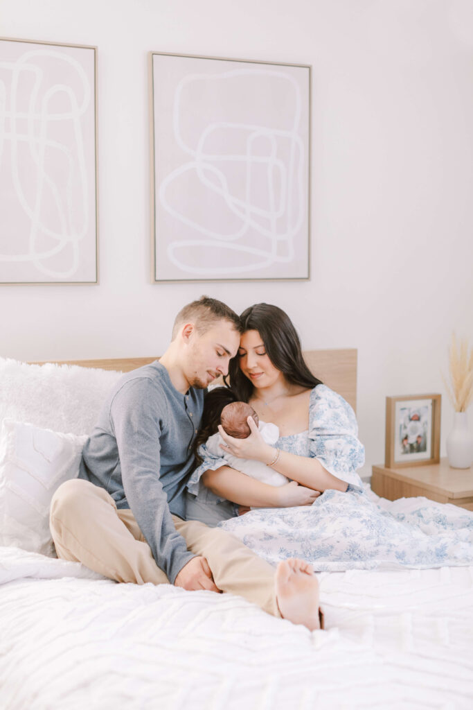 Image of Mom, Dad, and newborn sitting on a bed. The parents are snuggled together looking at their baby with the image being photographed from further away. This is an example of lifestyle family and newborn photography