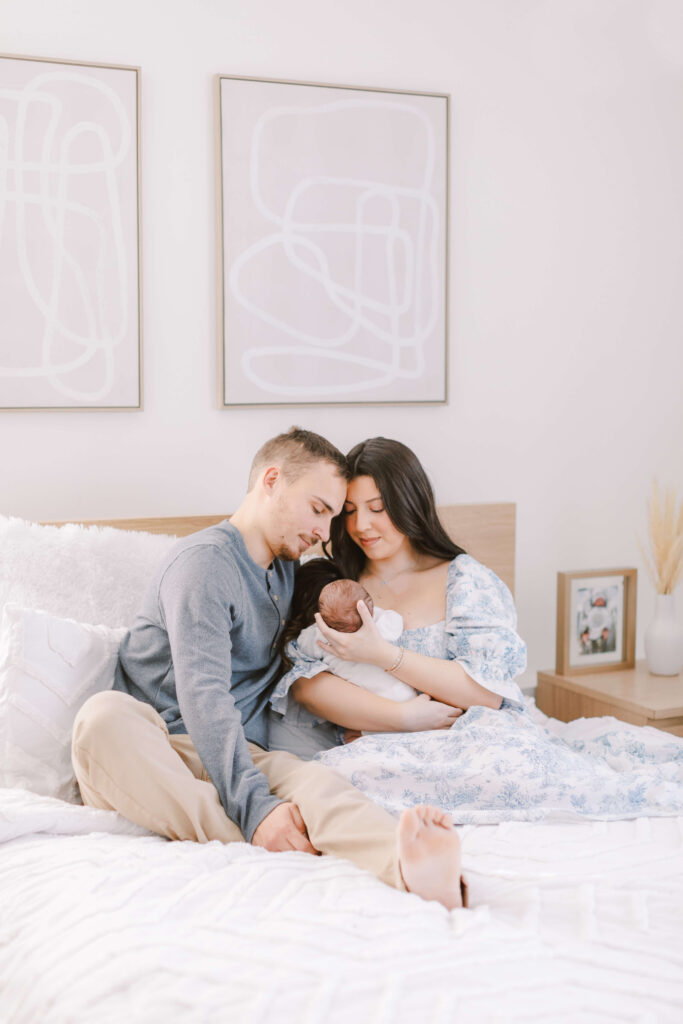 Choosing the Perfect Style Of Family and Newborn Photography For You Image of Mom, Dad, and newborn sitting on a bed. The parents are snuggled together looking at their baby with the image being photographed from further away. This is an example of lifestyle family and newborn photography