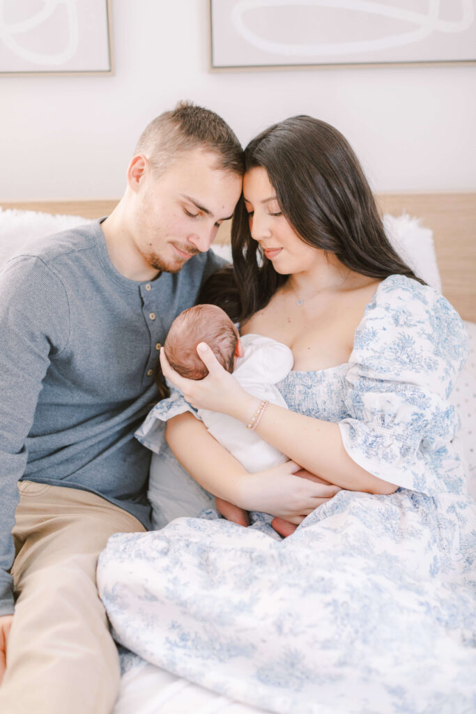 Image of Mom, Dad, and newborn sitting on a bed. The parents are snuggled together looking at their baby. This is an example of lifestyle family and newborn photography