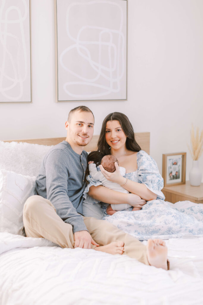 Image of Mom, Dad, and newborn sitting on a bed, smiling at the camera. This is an example of lifestyle family and newborn photography
