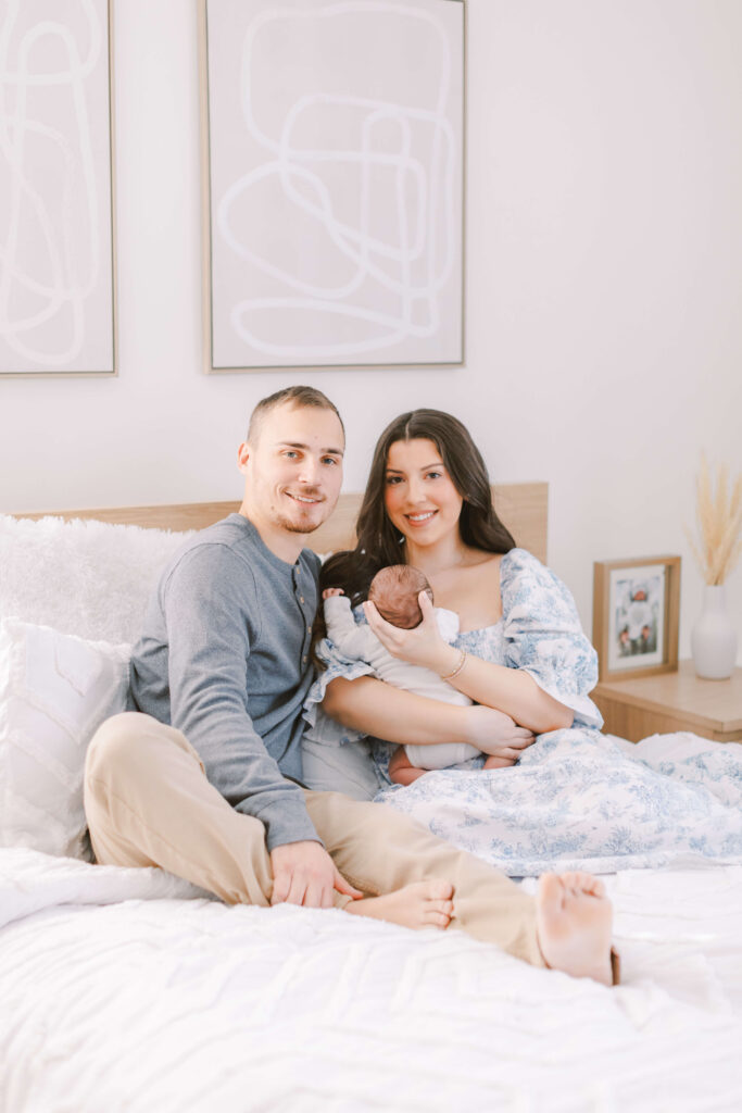 Choosing the Perfect Style Of Family and Newborn Photography For You Image of Mom, Dad, and newborn sitting on a bed, smiling at the camera. This is an example of lifestyle family and newborn photography