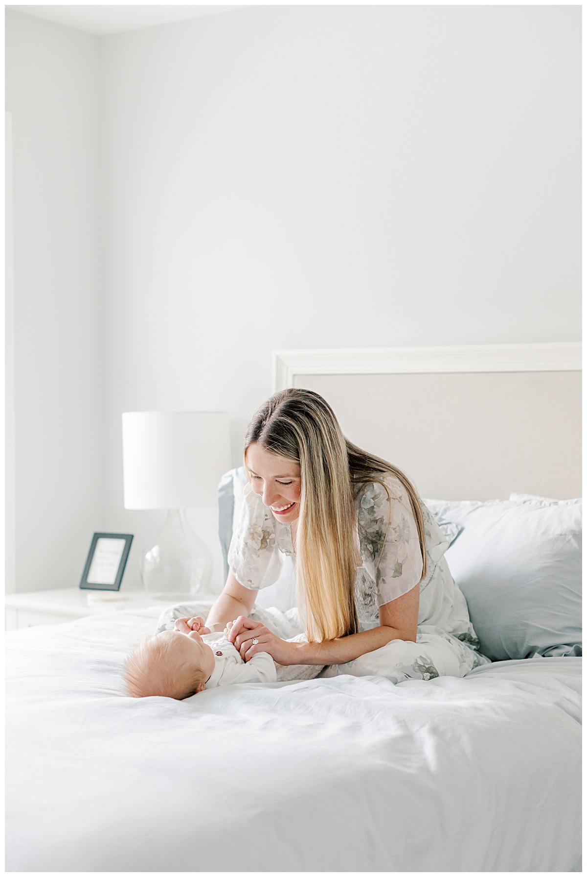 A new mom sits and stands by the window wearing a sage green floral dress. She is holding her newborn son during his in home newborn photo session.