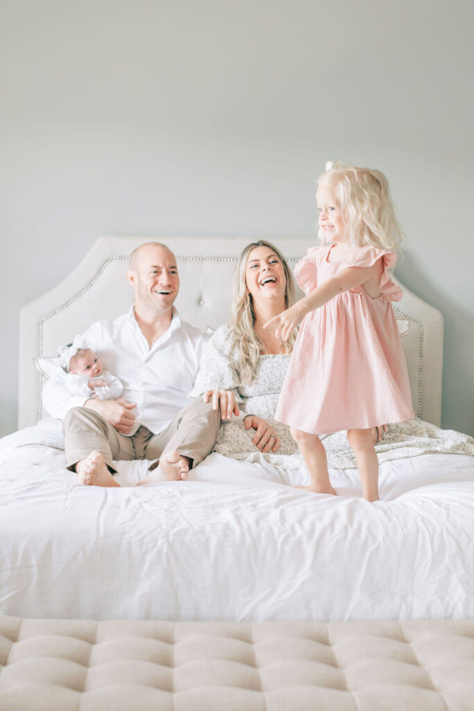 A family sits on the bed during their easy in-home newborn session and laughs while watching their toddler daughter dance