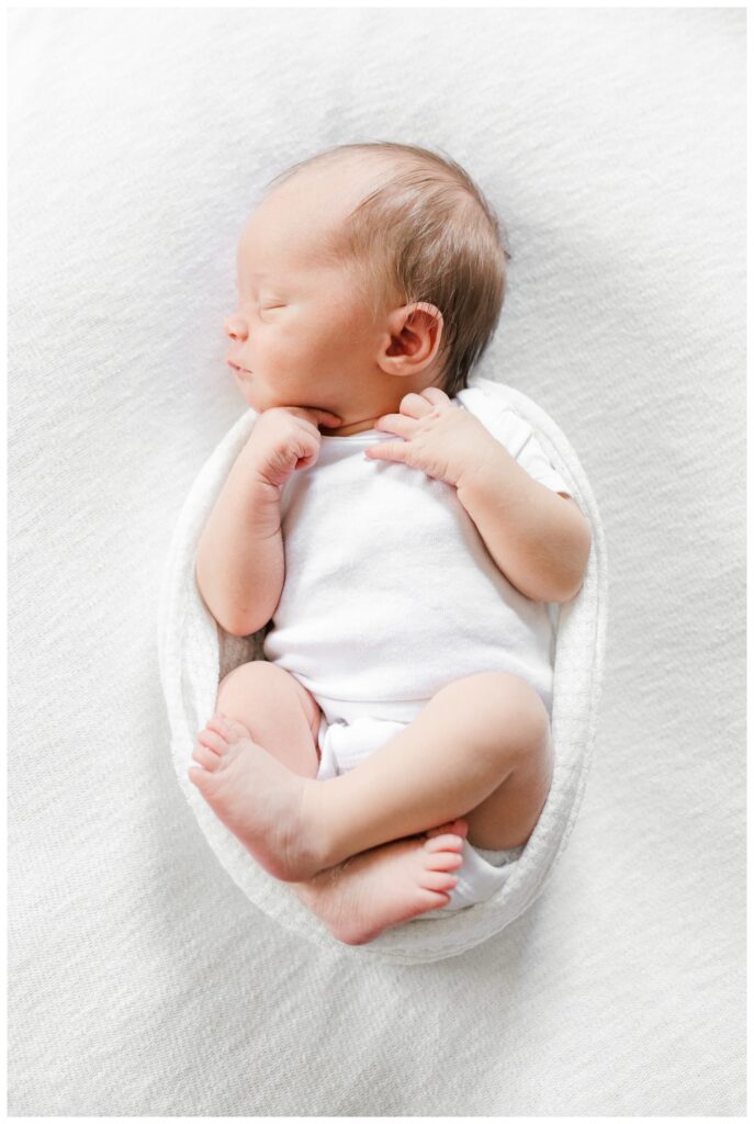 A newborn baby boy wearing a white onesie and wrapped in a white swaddle. His hands and feet are showing. HIs feet are crossed at the ankles and tucked up towards his stomach and his hands are up under his chin. This photo is being used as an example of when to take newborn photos at 5-10 days old.