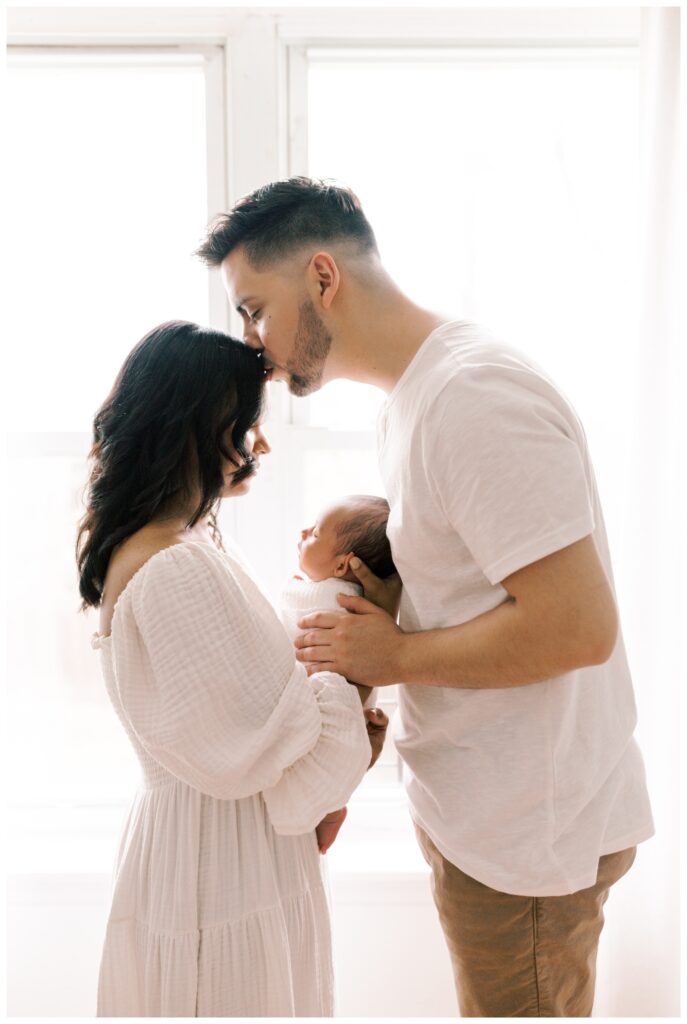 A couple stands facing eachother in front of a window during their lifestyle in-home newborn session. The mother is holding her newborn in front of her and the father is kissing the mother on her forehead. The mother is wearing a white dress and the father is wearing a white shirt and khaki pants.