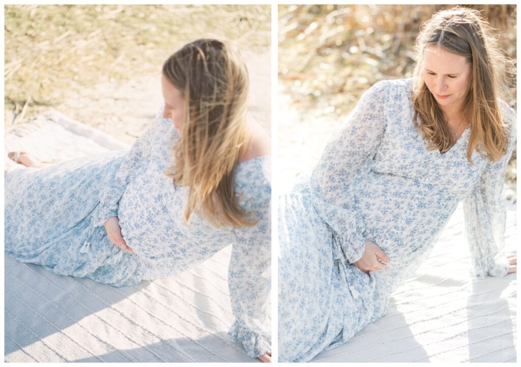A woman sits on a white blanket during her beach maternity photos. She is wearing a long sleeve white dress with blue flowers