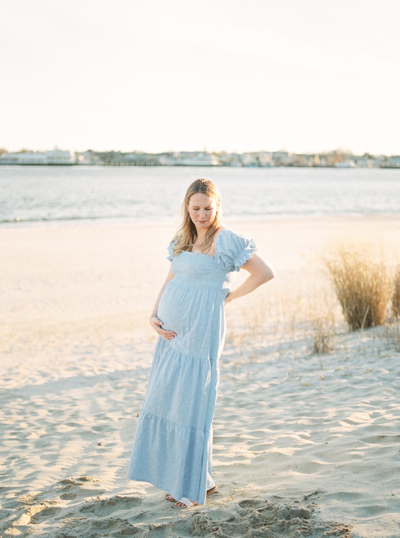 A woman wearing a long blue dress with short sleeves cradles her stomach during her beach maternity photos