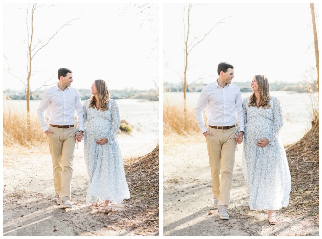 A couple holds hands and walks down a path during their beach maternity photos. The woman is wearing a long white dress with blue flowers.