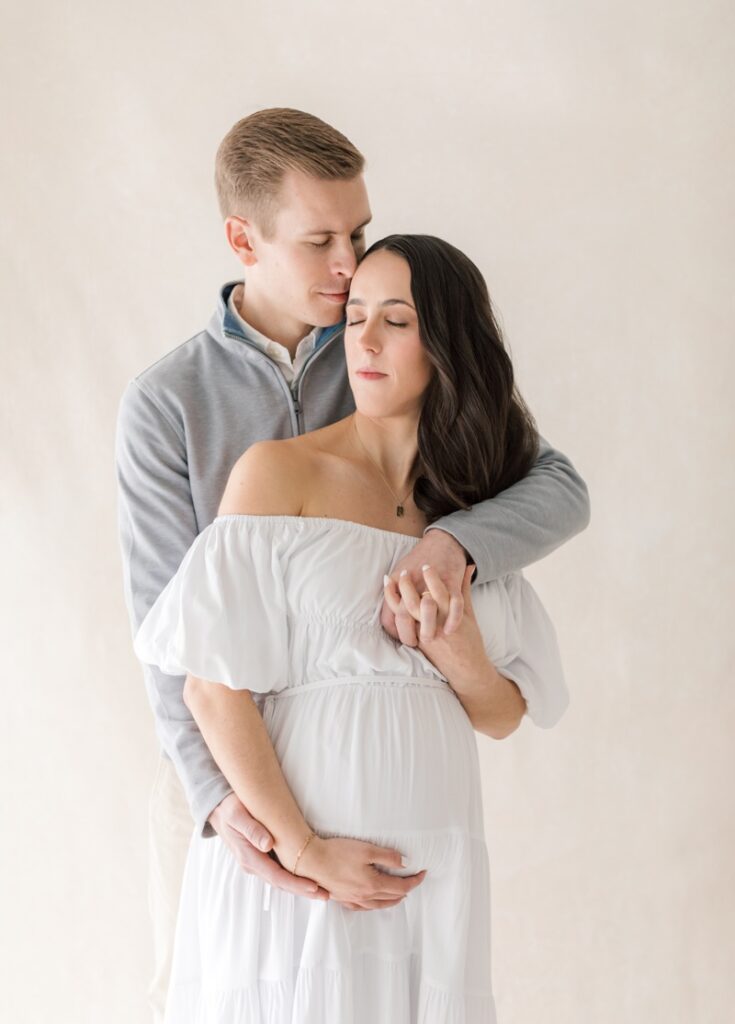 A couple snuggle together during studio maternity photos after loss. The mother is wearing a white dress.