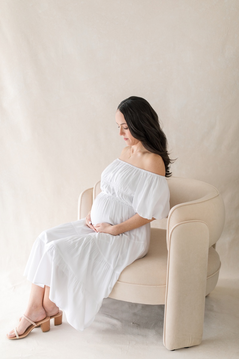 A pregnant woman sitting on a tan chair wearing a white dress during her maternity photos after loss