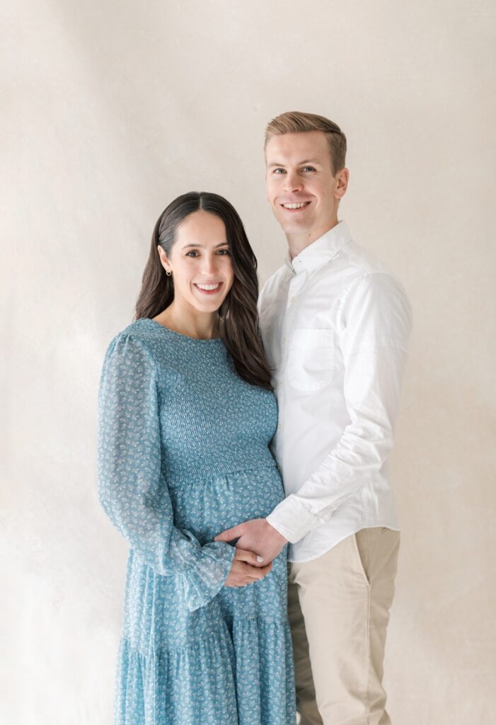 A couple smiles at the camera during their studio maternity photos after loss. The mother is wearing a blue floral dress.