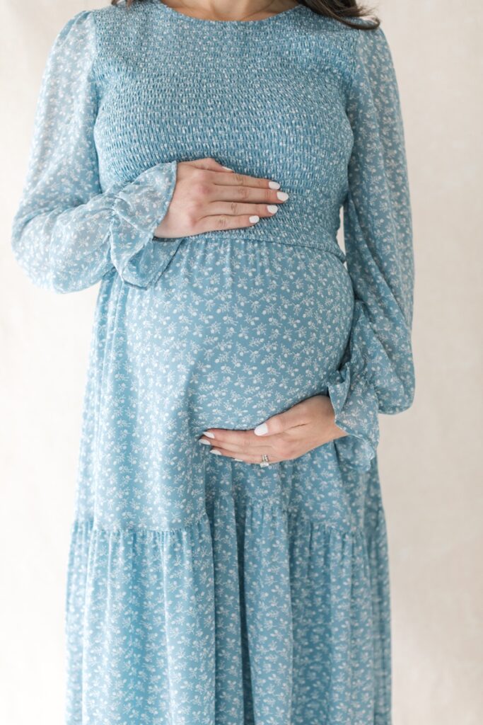 A pregnant woman wearing a  blue floral dress cradles her stomach during her studio maternity photos after loss