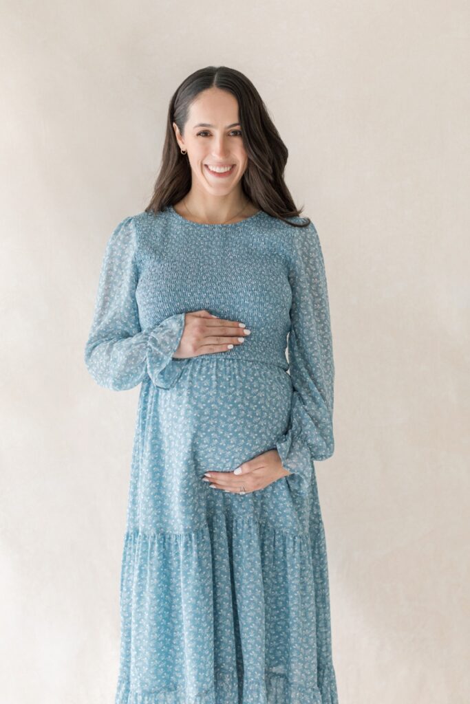 A pregnant woman wearing a blue floral dress cradles her stomach during her studio maternity photos after loss