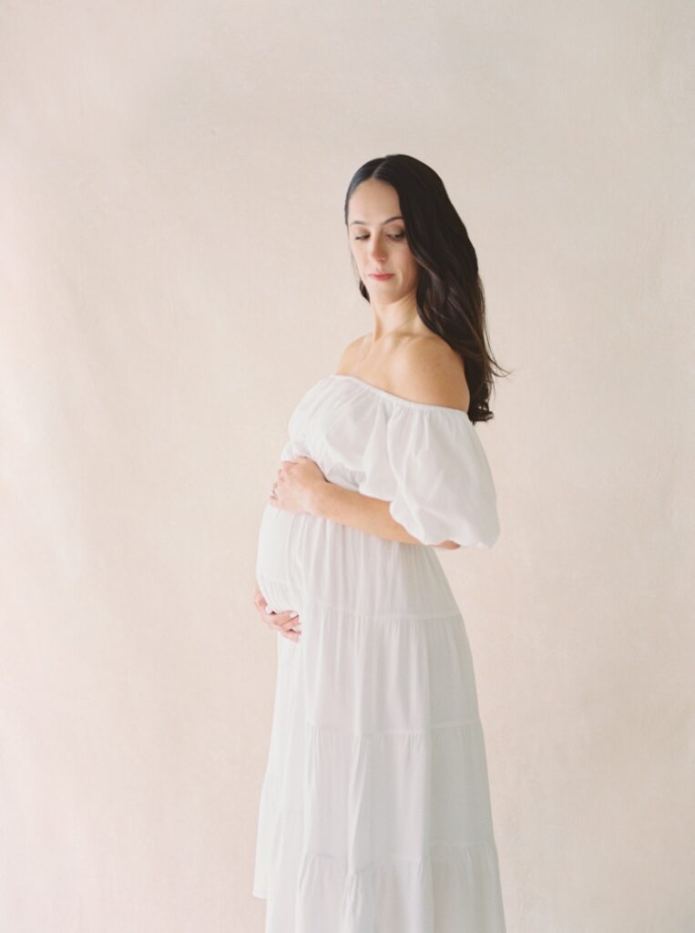 A pregnant woman wearing a white off the shoulder dress cradles her stomach during her studio maternity photos after loss