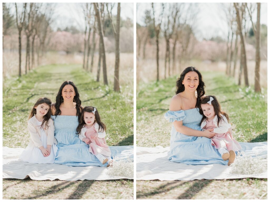 A mother in a blue dress sits with her daughters on a white blanket during their Mommy and Me photoshoot. 