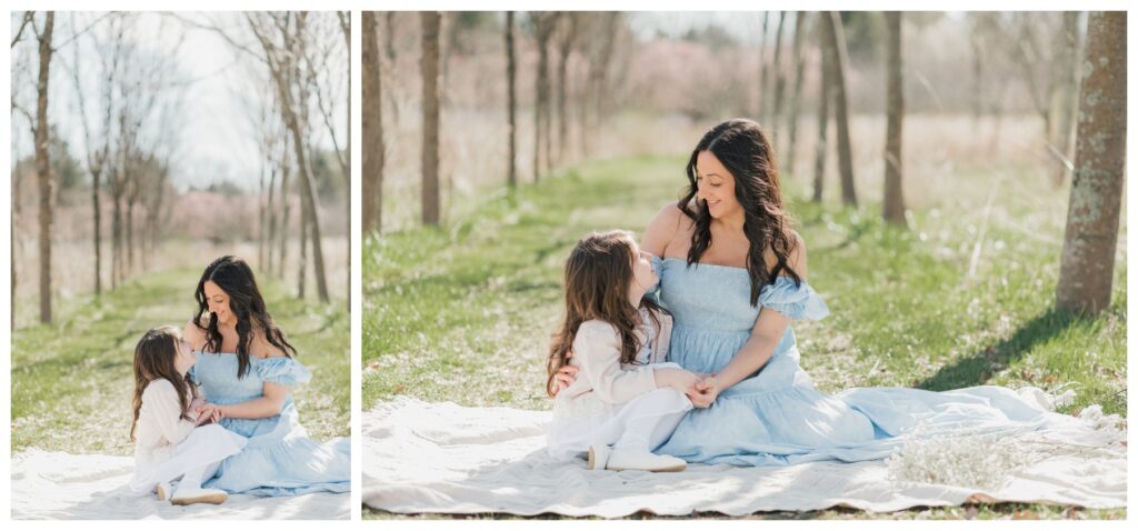 A mother in a blue dress sits with her daughter who is wearing a white dress. They are sitting on a white blanket. 