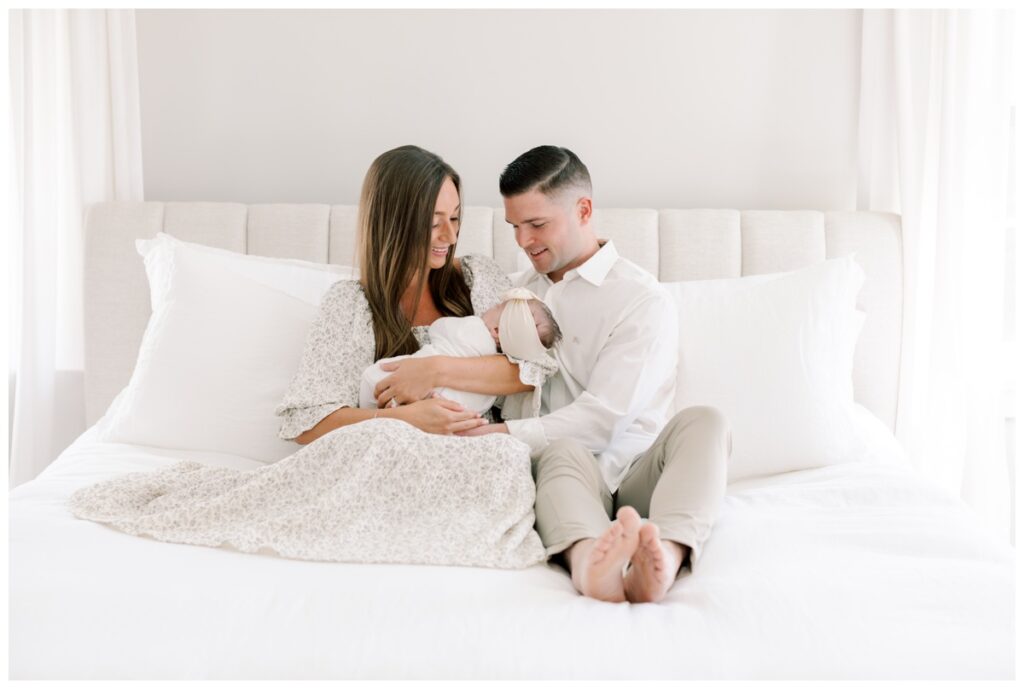 A couple sits close together on their bed holding their newborn baby girl and smiling down at her during their Cranbury NJ newborn photography session photographed by Kate Voda Photography