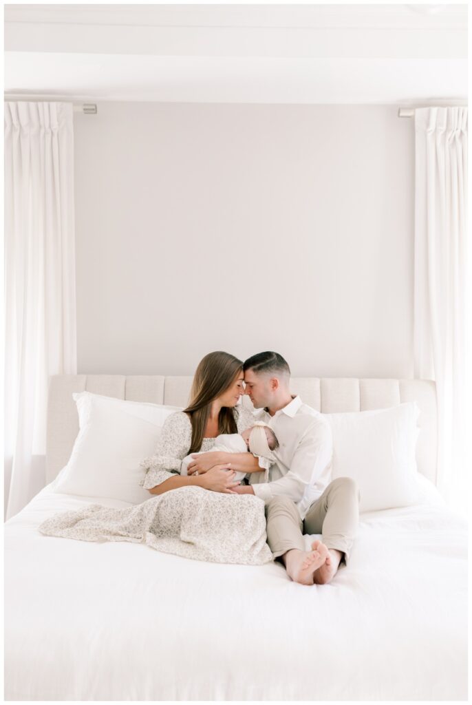 A couple sits close together on their bed holding their newborn baby girl between them during their newborn photos photographed by Kate Voda Photography