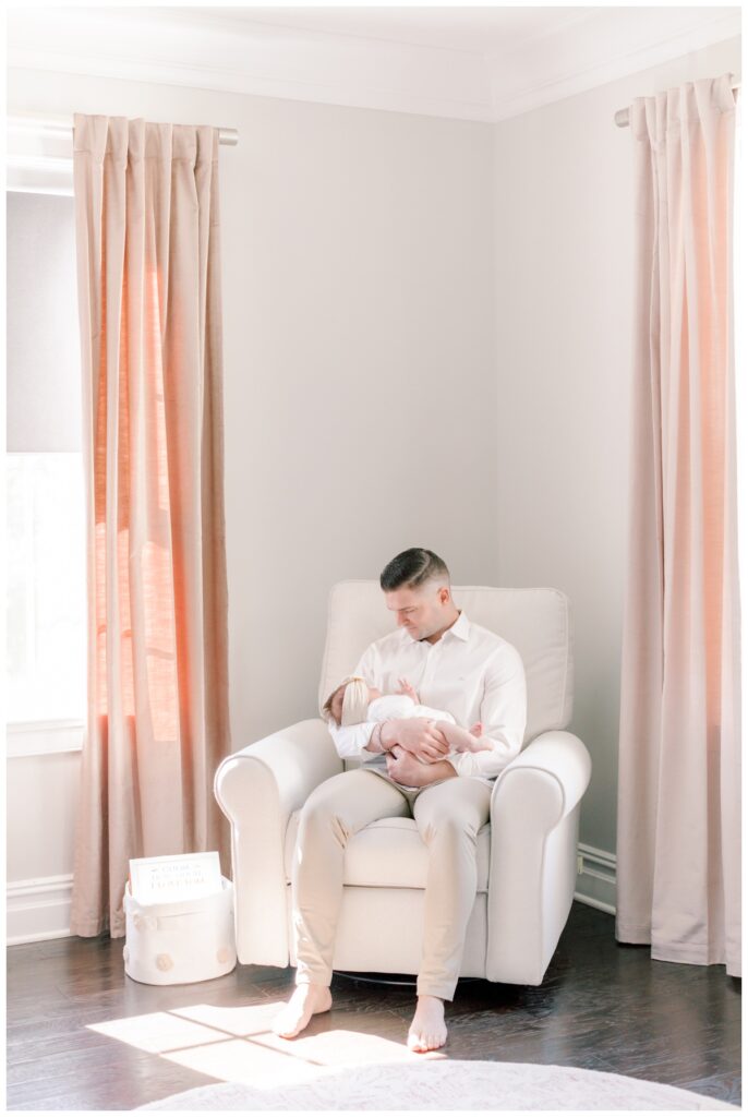 A father sits in the rocking chair in a newborn nursery while holding his baby girl.
