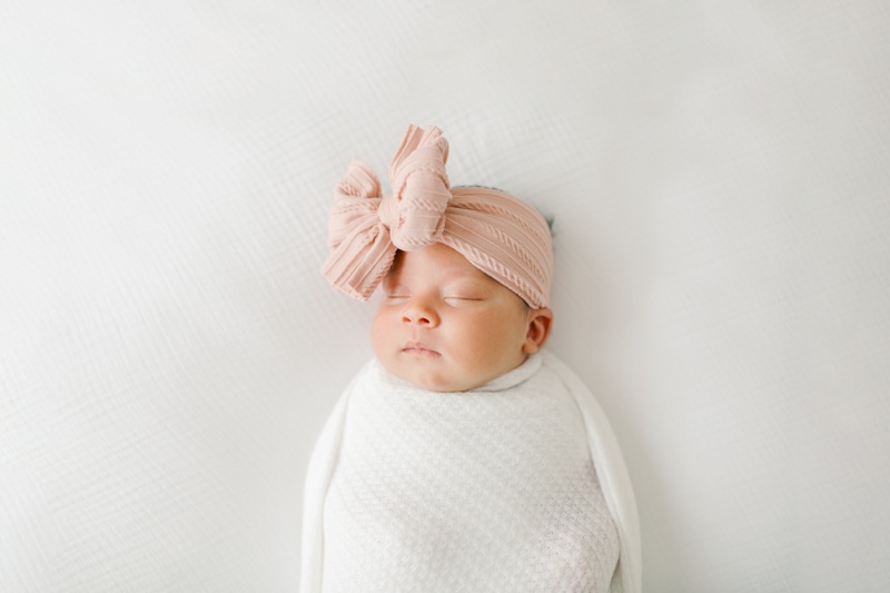 Newborn baby girl wrapped in a white swaddle with a large pink bow on her head, photographed by Kate Voda Photography