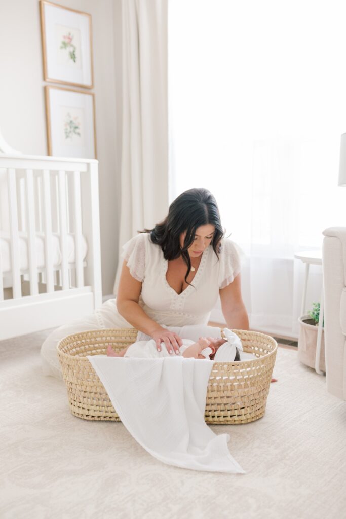 A mother sitting on the floor in the nursery looking down at her baby who is snuggled in a Moses basket during in-home newborn photos in New Jersey. Mom is wearing a white dress.