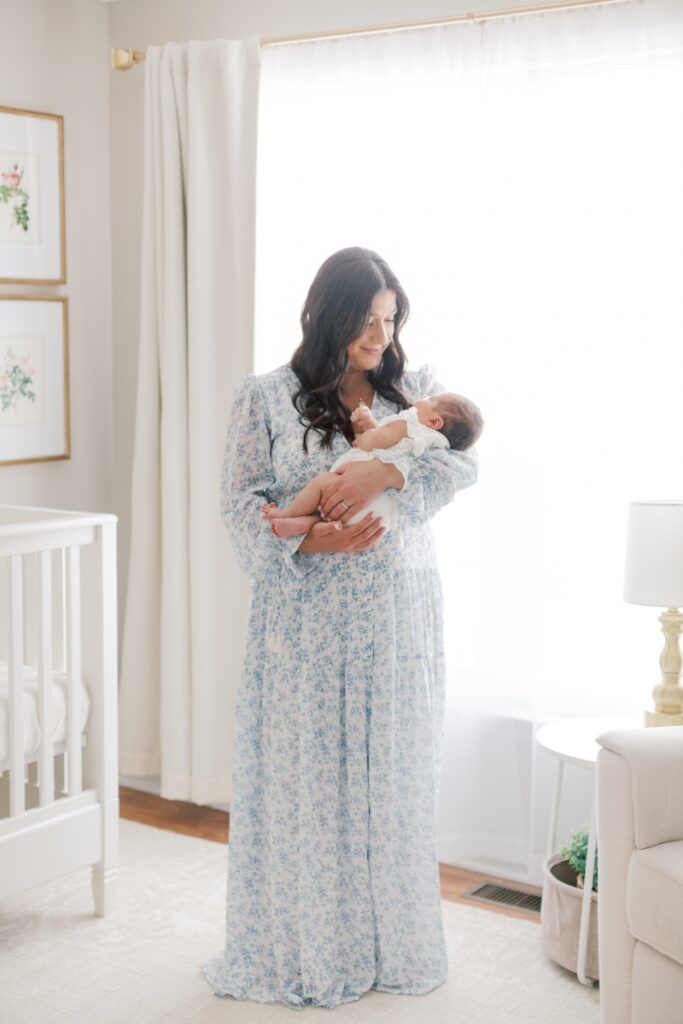 A full length photo of a mother looking down at her baby in her arms during in-home newborn photos in New Jersey. Mom is wearing a white dress with blue flowers on it.