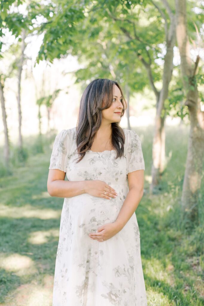 A pregnant woman in a floral dress cradles her belly while she stands on a tree covered pathway during outdoor maternity photos in New Jersey