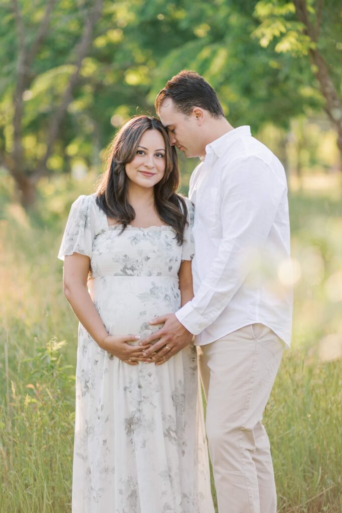 A couple stands close together during outdoor maternity photos in New Jersey. The woman is wearing a floral dress and looking at the camera while her husband rests his hand on her belly and his forehead on her head. 