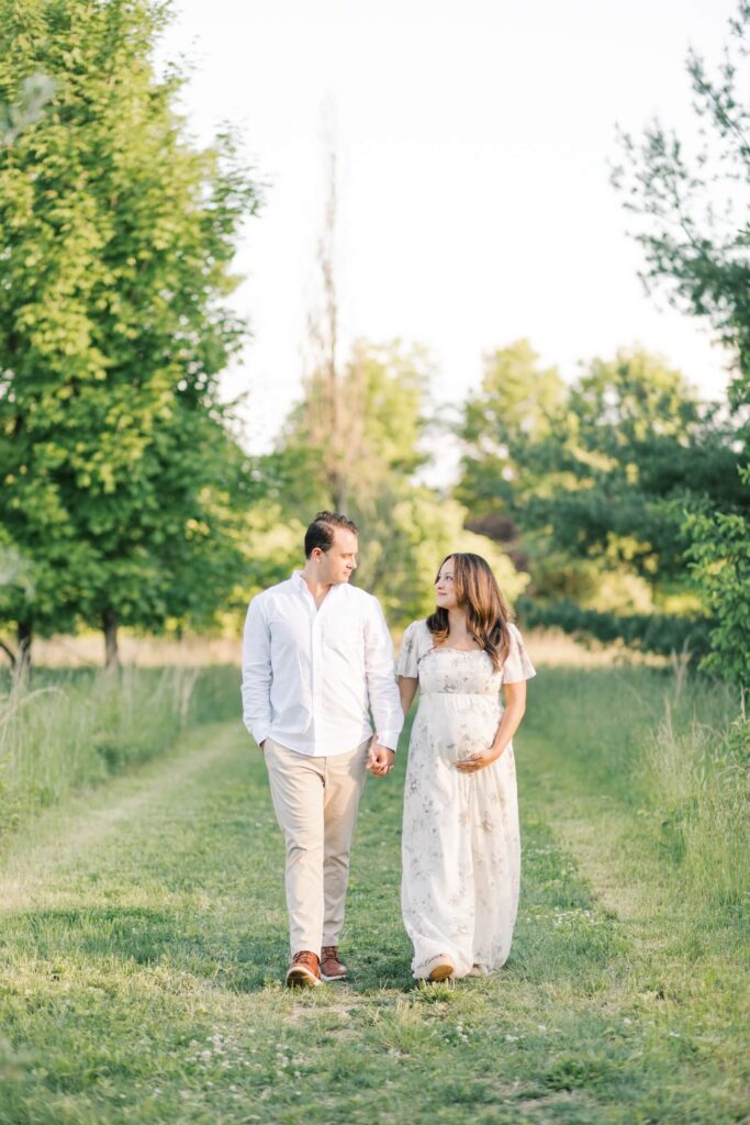 A couple holds hands and walks on a grassy pathway during outdoor maternity photos in NJ. 