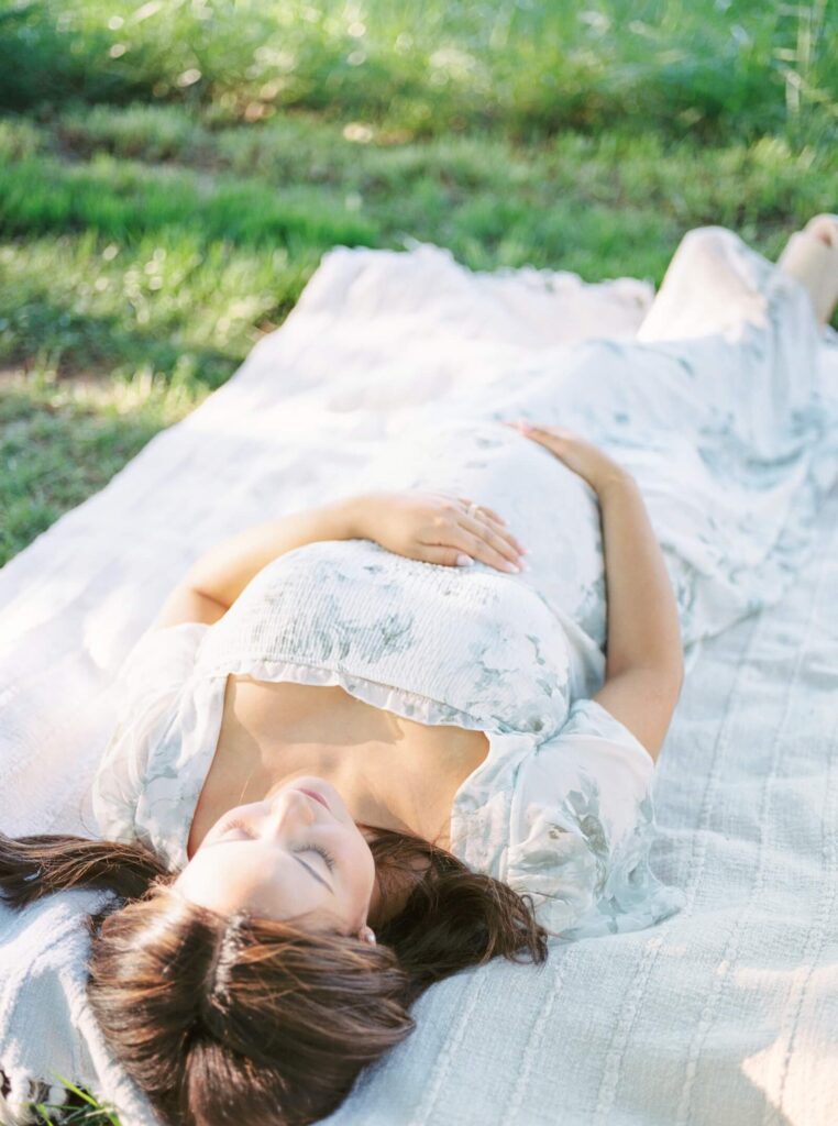 A pregnant woman lays on a blanket during her outdoor maternity photos in NJ. She is cradling her belly and there are beautiful pockets of sunlight all around her. 