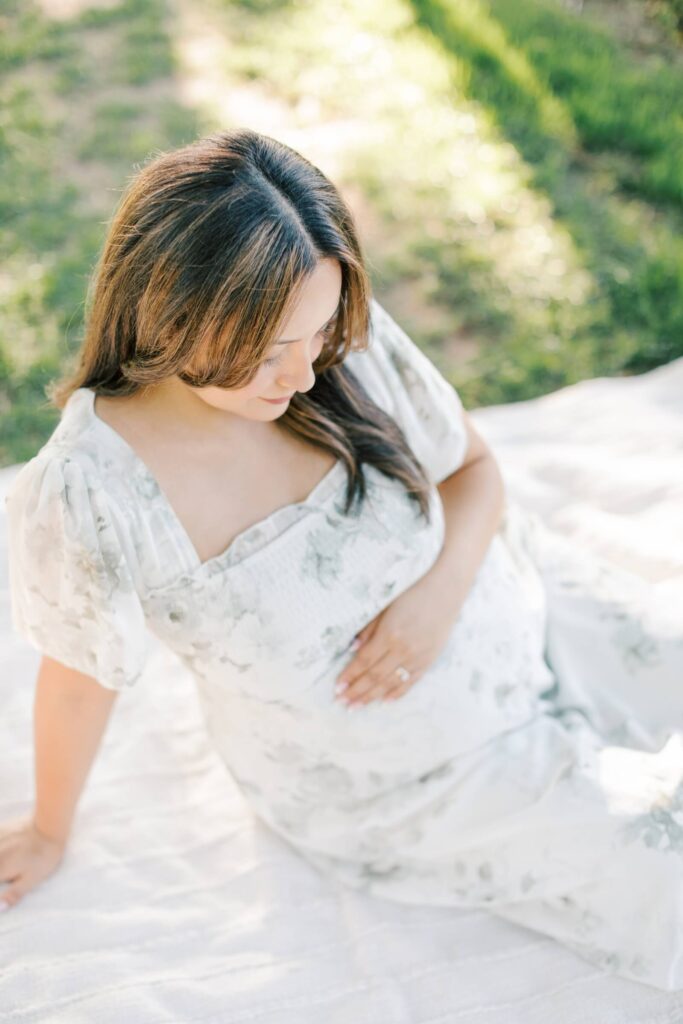 A pregnant woman sits on a blanket in the sun with her hand resting lightly on her belly. She is wearing a floral dress and is being photographed by NJ Maternity Photographer Kate Voda