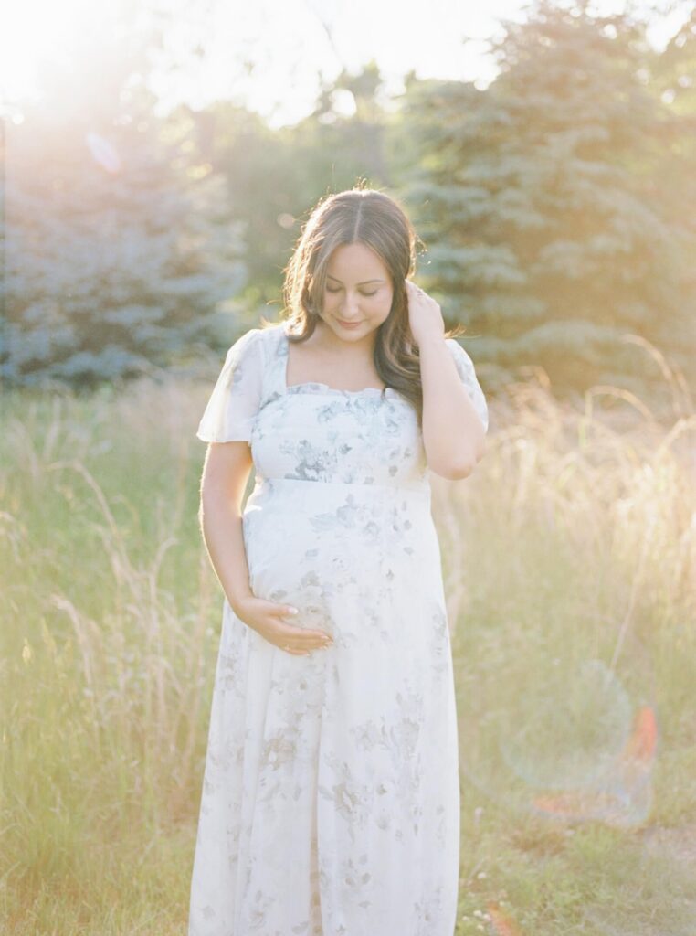 A pregnant womand holds her belly with one hand while she rests her other hand in her hair. She is wearing a floral dress and standing in front of tall grass during her outdoor maternity session photographed by Kate Voda Photography.