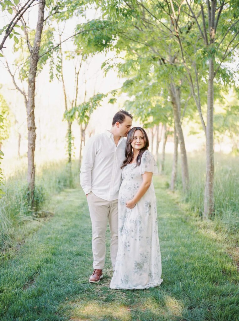 A mand and a pregnant woman in a floral dress stand close together on a tree covered pathway during outdoor maternity photos in New Jersey