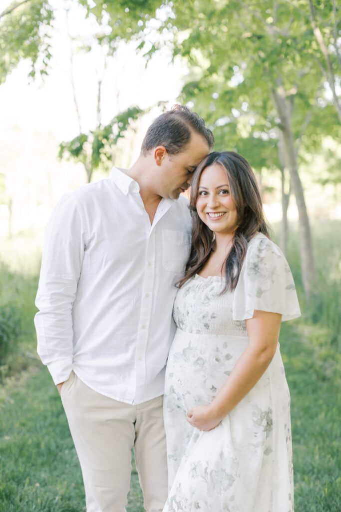 A man and a pregnant woman in a floral dress stand close together on a tree covered pathway during outdoor maternity photos in New Jersey