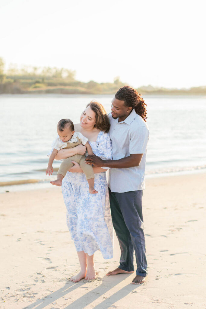 Mini Session, Half, or Full: Which Is Right for Your Family? (2026) Mom holding her toddler as dad hugs them both by the water's edge at Fisherman's Cove in Manasquan, NJ