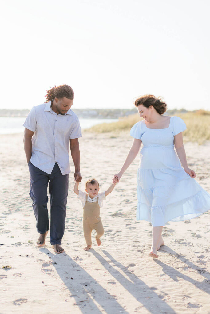 Mini Session, Half, or Full: Which Is Right for Your Family? (2026) Mom and dad walking barefoot on the sand, holding their toddler's hands as he takes steps between them at Fisherman's Cove