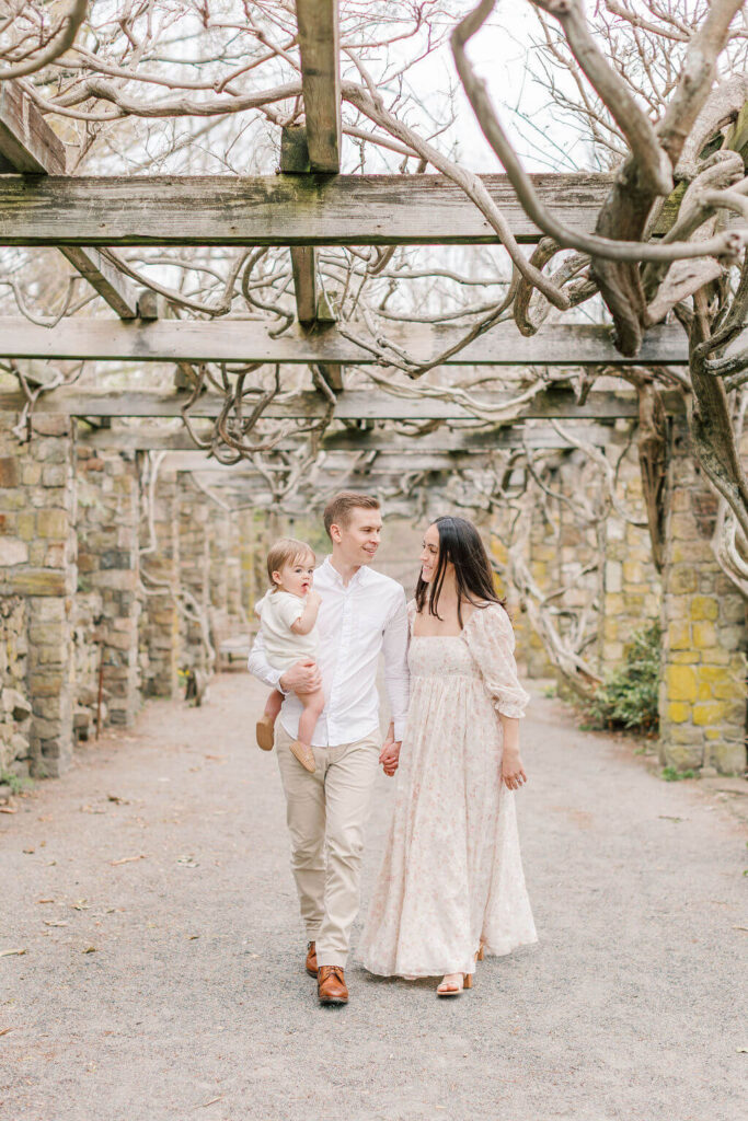 Mini Session, Half, or Full: Which Is Right for Your Family? (2026) Family walking hand in hand under the wisteria pergola at Cross Estate Gardens, with dad carrying their one year old