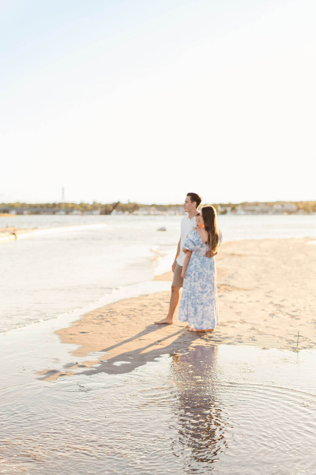Mom and dad standing together at the water's edge at Fisherman's Cove in Manasquan, NJ, with their toddler in mom's arms and their reflection in the tide pool