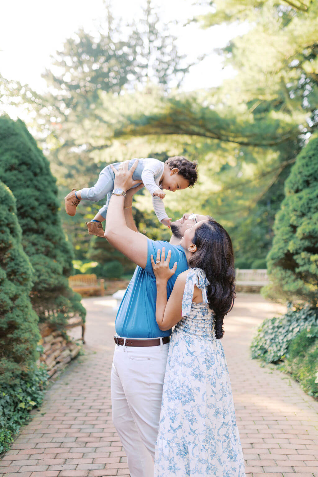Dad lifting a smiling toddler overhead as mom hugs him, surrounded by evergreens at Sayen House and Gardens
