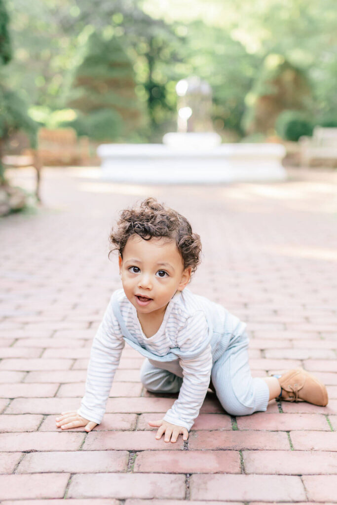 Mini Session, Half, or Full: Which Is Right for Your Family? (2026) Curly haired toddler crawling on a brick path and looking up at the camera, with the garden fountain behind him at Sayen House and Gardens