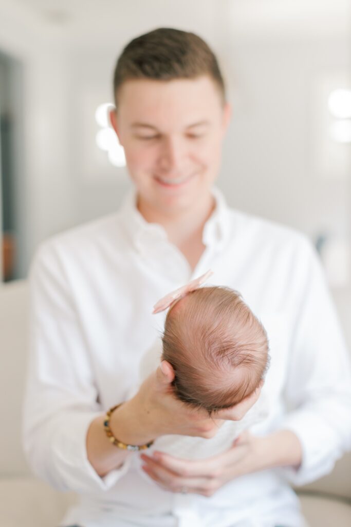 a man in a white shirt sits on the couch holding his newborn during a New Jersey in-home newborn photography session