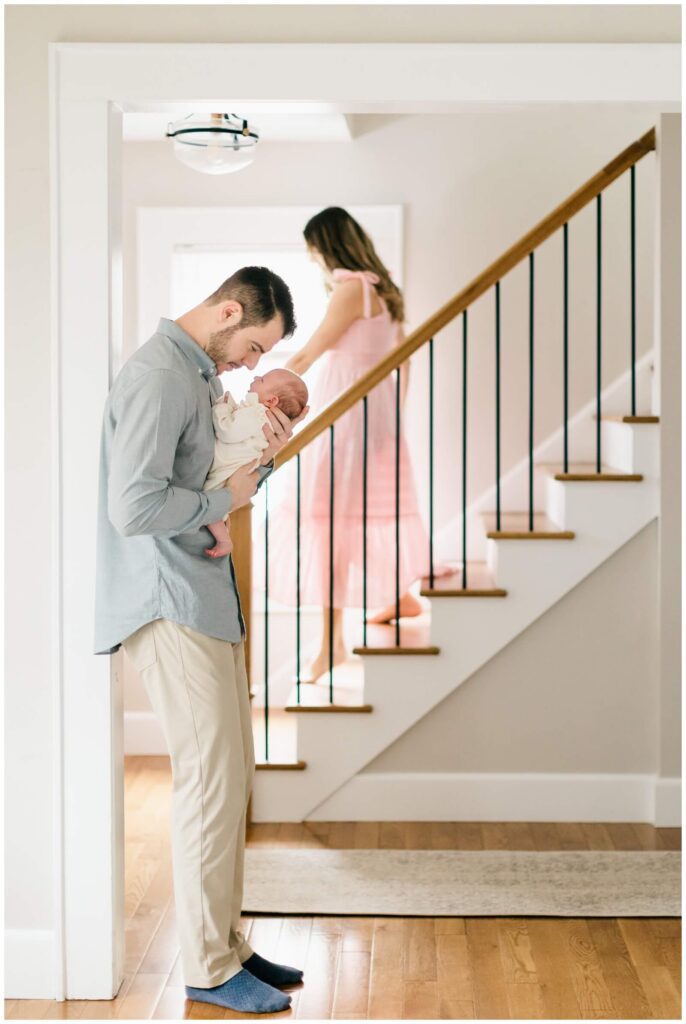 Candid family portraits in New Jersey — dad cradling newborn as mom comes down the stairs during lifestyle newborn session