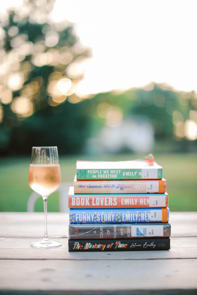 Stack of summer books next to a glass of rosé on an outdoor table at golden hour
