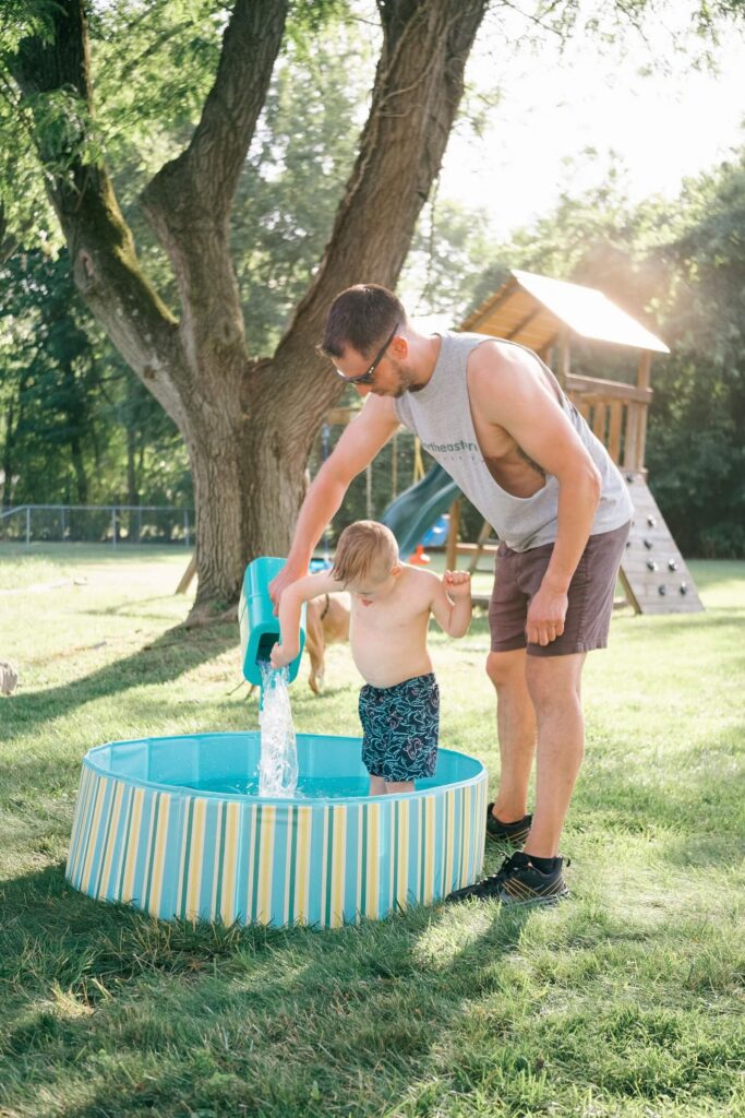Your August Photo Scavenger Hunt A dad pouring water with a toddler in a backyard kiddie pool on a summer afternoon — a photo scavenger hunt moment
