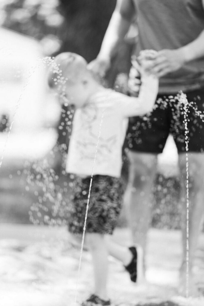 Your August Photo Scavenger Hunt Black and white photo of a child and parent at a backyard splash pad, shot through the water jets as part of a summer photo scavenger hunt